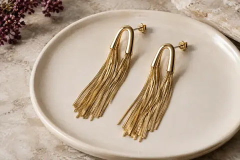 Paire de boucles d’oreilles dorées à longues franges, posée sur un plateau en céramique ivoire