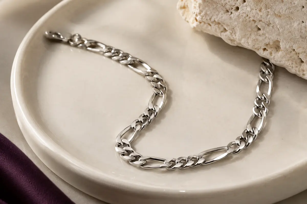 Bracelet chaîne Figaro argenté posé sur une céramique ivoire, avec ses maillons brillants visibles en gros plan.