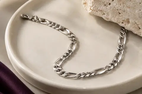 Bracelet chaîne Figaro argenté posé sur une céramique ivoire, avec ses maillons brillants visibles en gros plan.