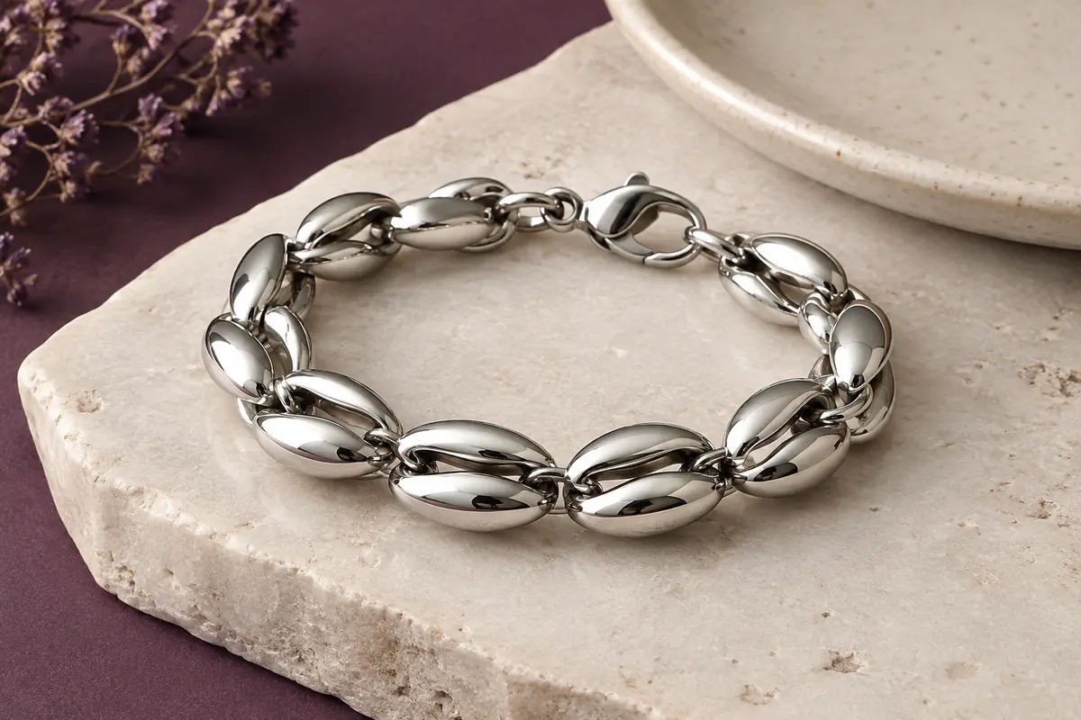 Bracelet argenté à maillons grain de café posé sur une pierre claire près d’un plateau en céramique.
