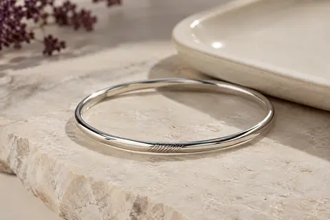 Bracelet jonc fin argenté à message gravé, posé sur une pierre claire près d’un plateau en céramique.