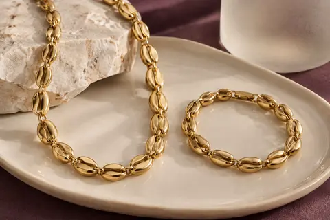 Collier et bracelet dorés à maillons grain de café disposés sur un plateau en céramique ivoire.