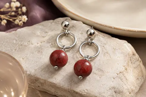 Boucles pendantes en acier argenté avec pierres naturelles rouges, posées sur une céramique ivoire.
