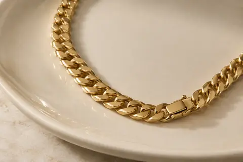 Collier gourmette cubain doré à larges maillons, disposé en courbe sur un plateau en céramique ivoire.