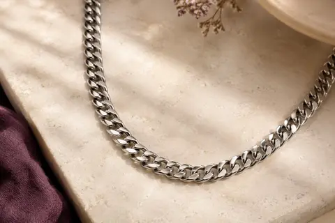 Collier ChainsHouse argenté à maillons cubains de 6 mm disposé en courbe sur une pierre claire.