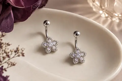Deux piercings de nombril argentés à motifs floraux, posés sur un plateau en céramique ivoire.