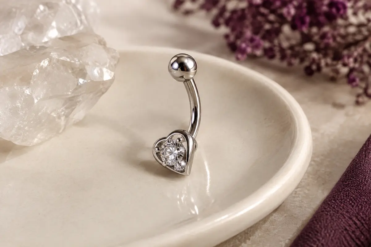 Piercing de nombril argenté à barre incurvée, avec un motif cœur et deux zircones claires.