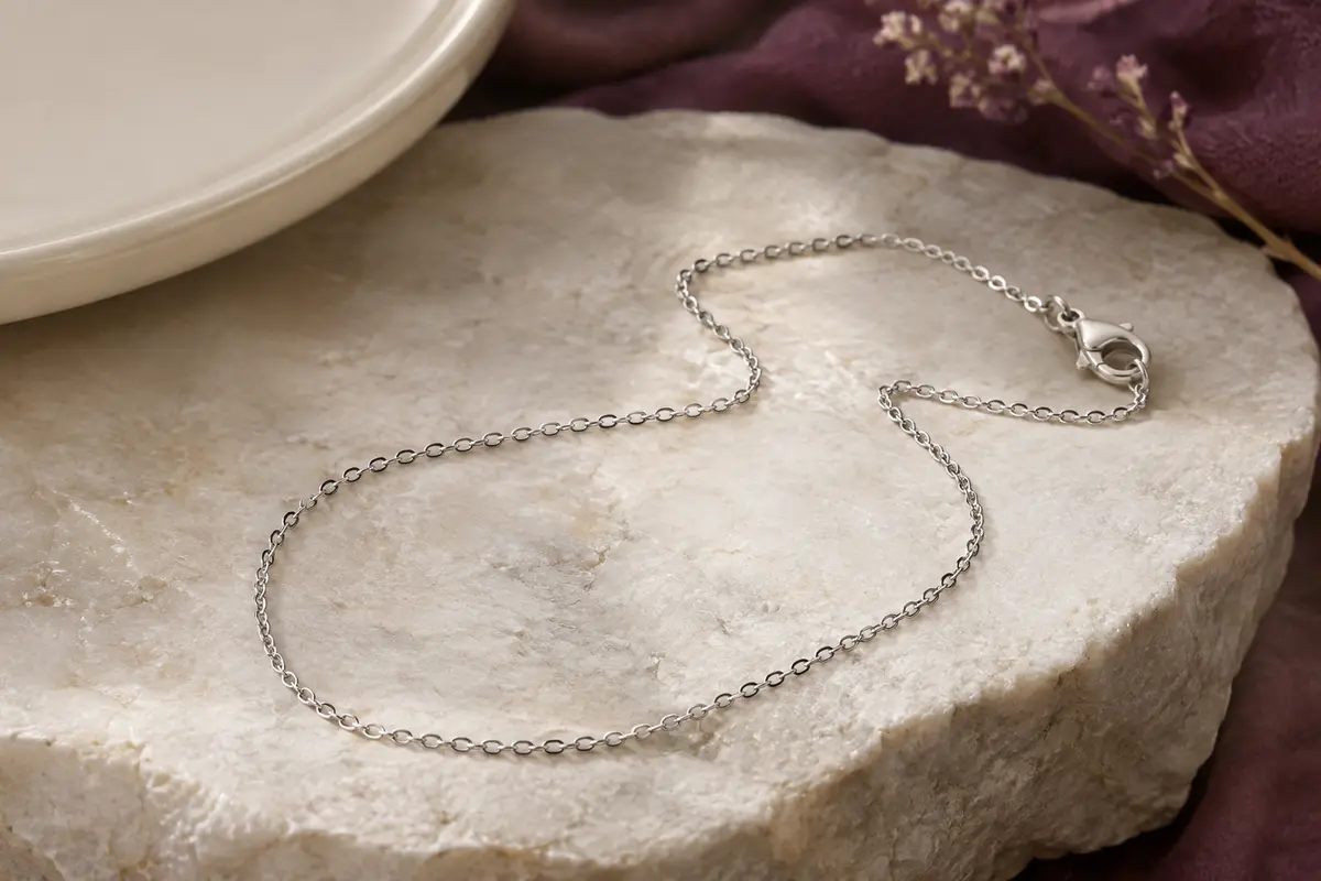 Collier argenté à fine maille forçat posé sur une pierre claire, avec son fermoir mousqueton visible