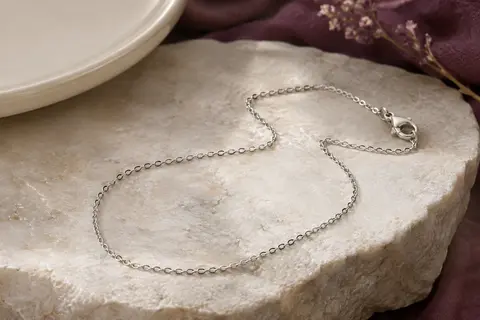 Collier argenté à fine maille forçat posé sur une pierre claire, avec son fermoir mousqueton visible