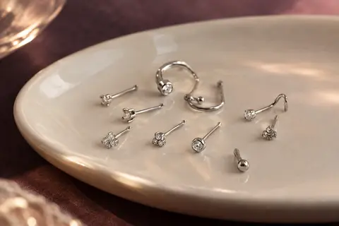 Lot de piercings de nez argentés avec de petits embouts CZ, disposé sur un plateau en céramique ivoire.