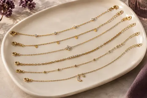 Six chaînes de cheville dorées ornées de zircons, disposées en accumulation sur un plateau en céramique claire.