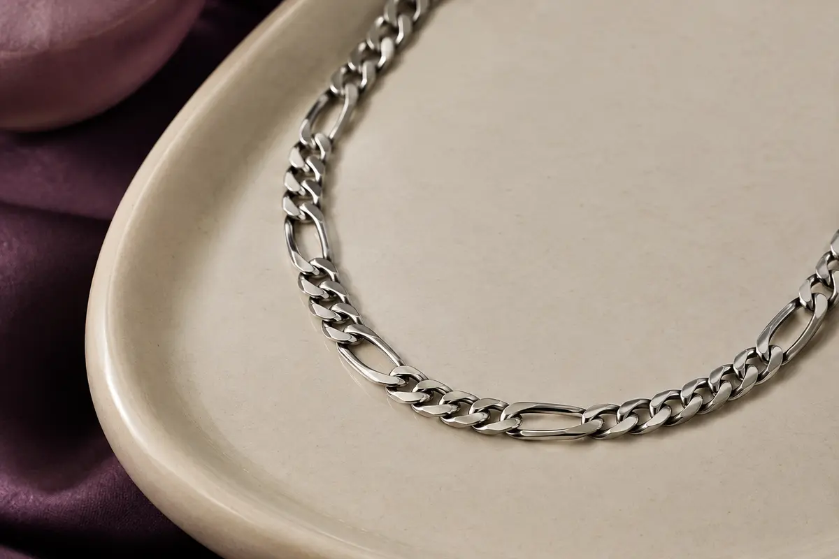 Collier Figaro argenté en acier inoxydable, disposé en courbe sur un plateau en céramique claire.