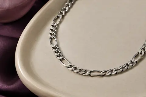 Collier Figaro argenté en acier inoxydable, disposé en courbe sur un plateau en céramique claire.