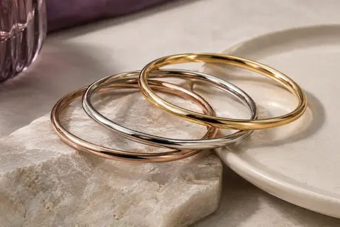 Trois joncs fins à finition dorée disposés en accumulation sur une pierre claire et une céramique ivoire.