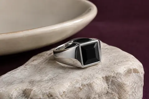 Chevalière argentée en acier poli avec une pierre noire carrée, posée sur une surface claire.