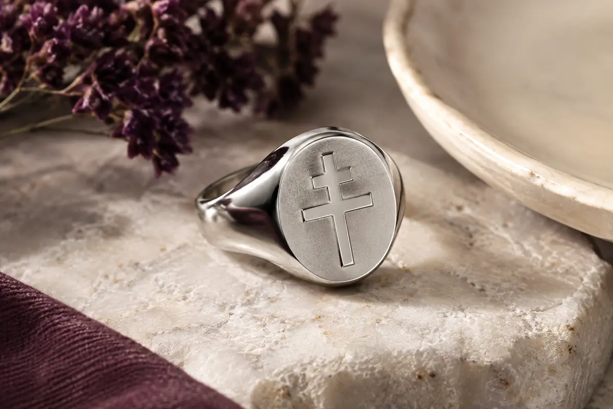 Chevalière en acier argenté ornée d’une croix de Lorraine, posée sur une pierre claire.