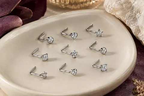 Neuf piercings de narine argentés en forme de L, ornés de zircons aux motifs variés sur une céramique claire.