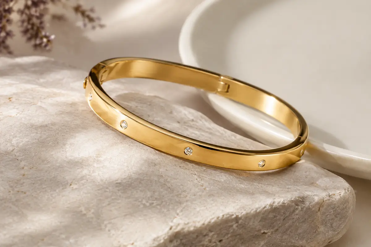 Bracelet jonc ovale doré avec détails scintillants et fermoir dissimulé, posé sur une pierre claire.