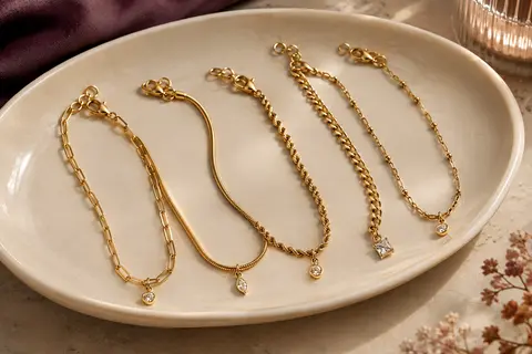 Cinq chaînes de cheville dorées aux maillons variés, disposées sur un plateau en céramique claire.