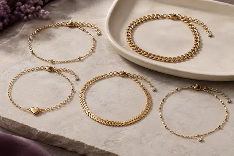 Cinq bracelets de cheville dorés aux mailles variées, disposés sur une pierre claire près d’un plateau en céramique.