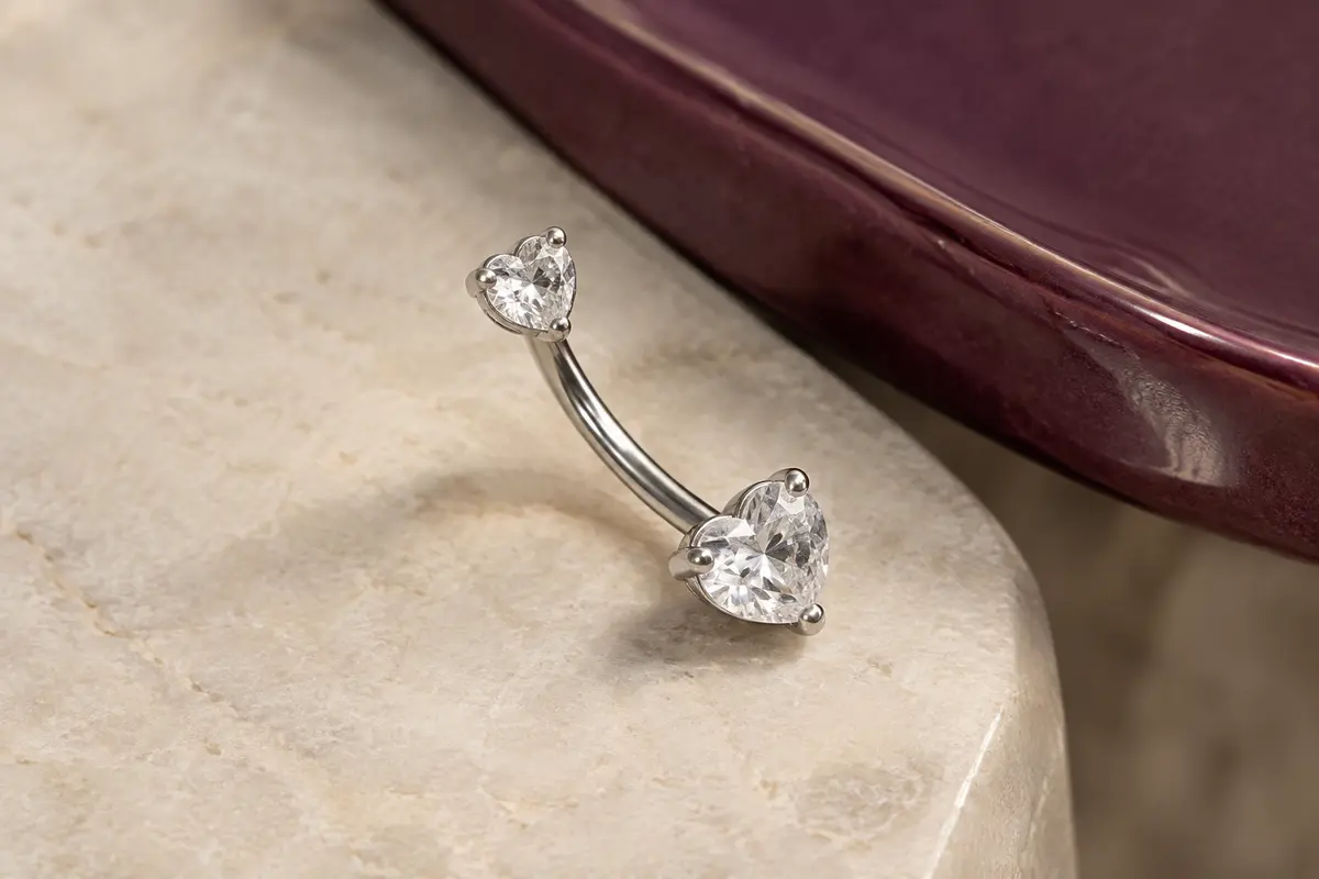 Piercing de nombril argenté à deux cœurs sertis de zircons transparents, posé sur une pierre claire.