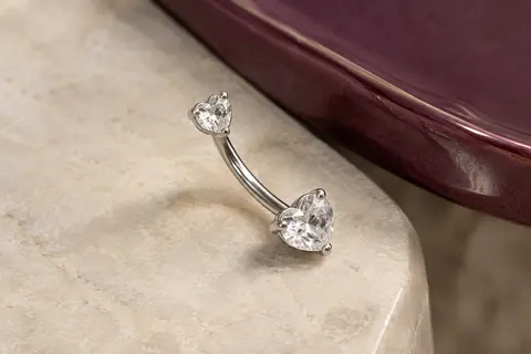 Piercing de nombril argenté à deux cœurs sertis de zircons transparents, posé sur une pierre claire.