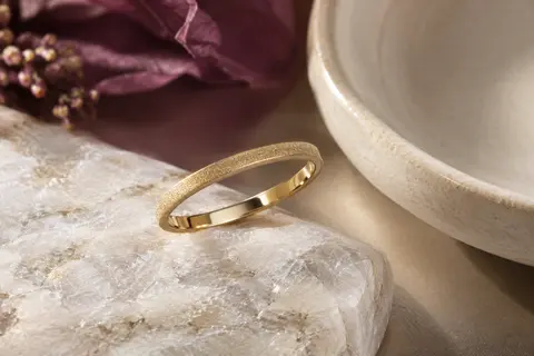 Bague alliance fine plaquée or à surface sablée, posée sur une pierre claire dans une lumière douce.