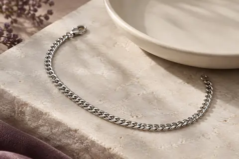 Bracelet chaîne argenté à maille cubaine fine, posé en courbe sur une pierre claire.