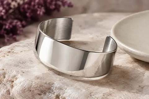 Bracelet manchette ouvert en acier argenté, large et poli, posé sur une pierre claire.