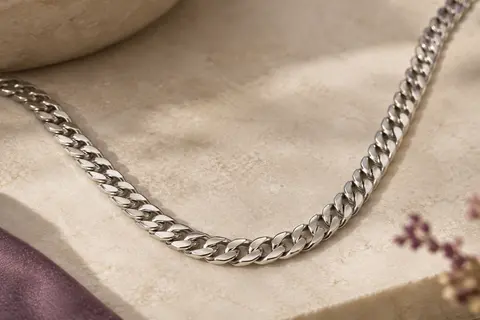 Collier Prosteel argenté à maille cubaine, disposé en courbe sur une pierre claire.