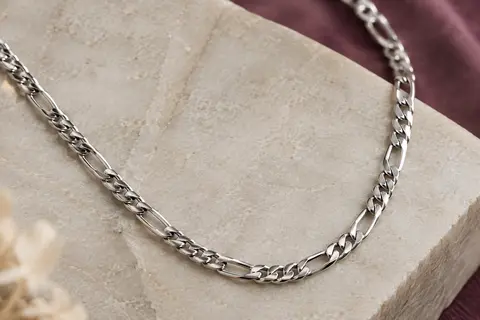 Collier Figaro argenté de 55 cm posé sur une pierre claire, avec ses maillons alternés visibles.