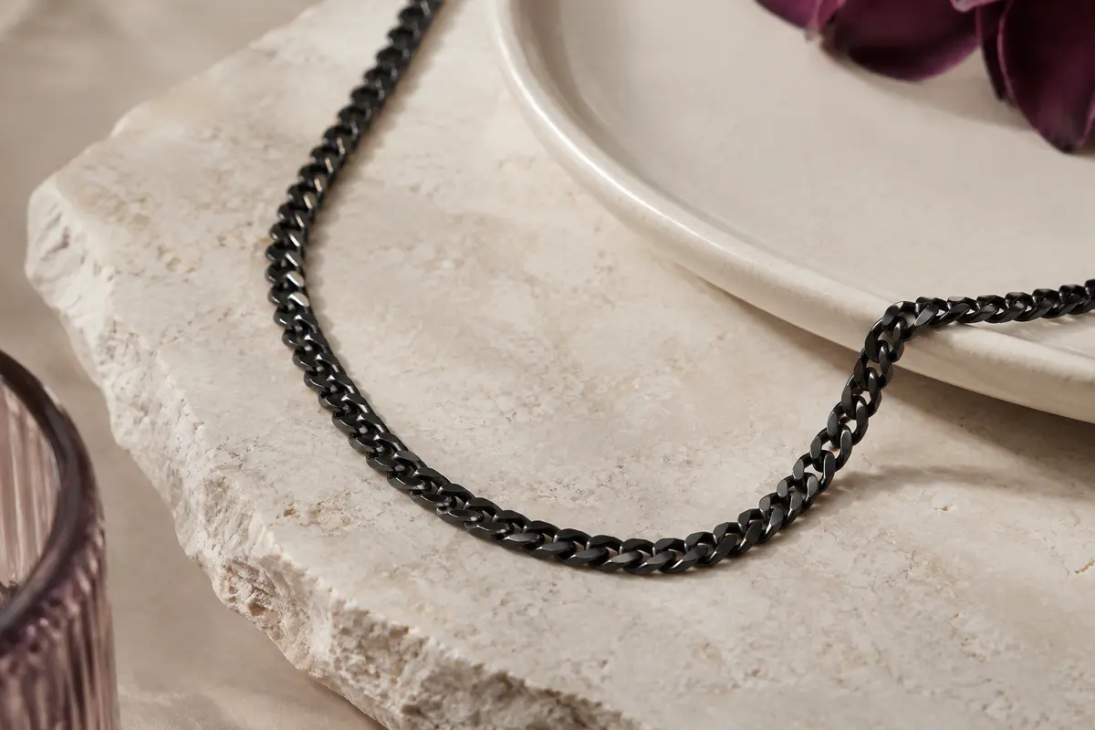 Collier chaîne cubaine noire à maillons de 4 mm posé sur une pierre claire près d’un plateau en céramique.
