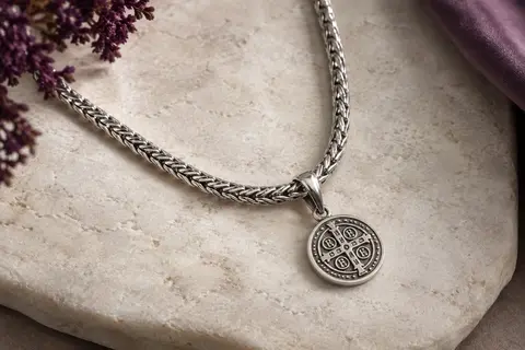 Collier argenté à chaîne spiga et pendentif rond orné d’une croix de Saint Benoît, posé sur une pierre claire.